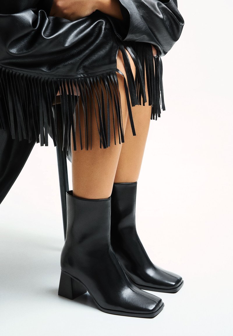 Stivaletti neri alla caviglia con punta quadrata e tacco a blocco, realizzati in pelle lucida, abbinati a un capo in pelle nera con frange.