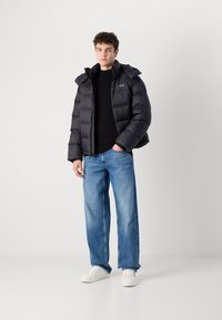 Must must jacket kapuutsiga, millel on tikitud disain, lukuga sulgemine ja liibuv siluett, koos siniste teksade ja valgete tossudega.