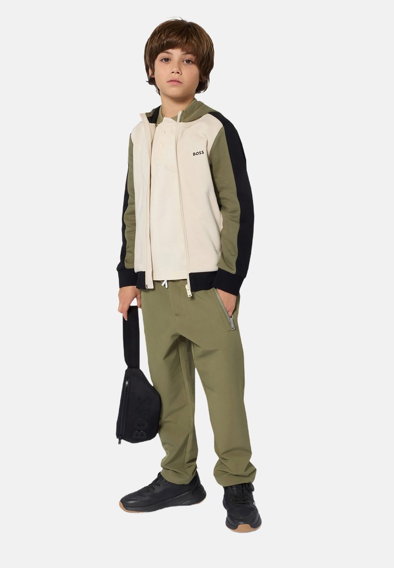 Veste zippée bicolore en crème et olive avec des manches noires, associée à un pantalon olive et des baskets noires. Petit logo sur la veste.