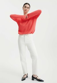 Korallfarbener Strickpullover mit weiten Ärmeln, kombiniert mit weißen, geraden Jeans und schwarzen Blockabsatzschuhen, vor einem hellen Hintergrund.