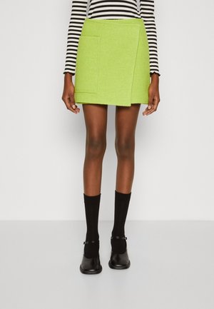 Kobieta ubrana w mini spódniczkę w kolorze jasnozielonym z przednią kieszenią, czarne skarpetki, czarne buty Mary Jane oraz czarno-biały, paskowany top z długim rękawem.