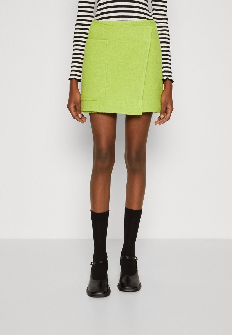 Femme portant une mini-jupe vert citron avec une poche avant, des chaussettes noires, des chaussures Mary Jane noires et un haut à manches longues rayé noir et blanc.