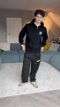 Fekete cipzáros kapucnis pulóver logóval, fekete melegítőnadrággal, amelynek oldalsávjai vannak. Szürke sportcipővel viselve egy texturált fehér szőnyegen.