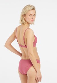 Donna bionda in bikini rosa a righe guarda oltre la spalla contro uno sfondo bianco, con una mano appoggiata sui fianchi.