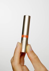 Prodotto labbra BareMinerals in un cilindro metallico oro con una fascia arancione, caratterizzato da una texture liscia e da una finestra trasparente per visualizzare la tonalità.
