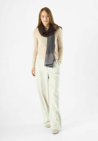 Sciarpa marrone e grigia realizzata in tessuto morbido, drappeggiata in modo casual. Texture liscia con bordi frangiati. Abbinata a pantaloni e top di colore chiaro.
