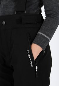 Salopettes noires imperméables avec bretelles réglables, poches zippées sur les côtés et détail de marque sur la cuisse. Comprend un poignet de manche en gris doux.