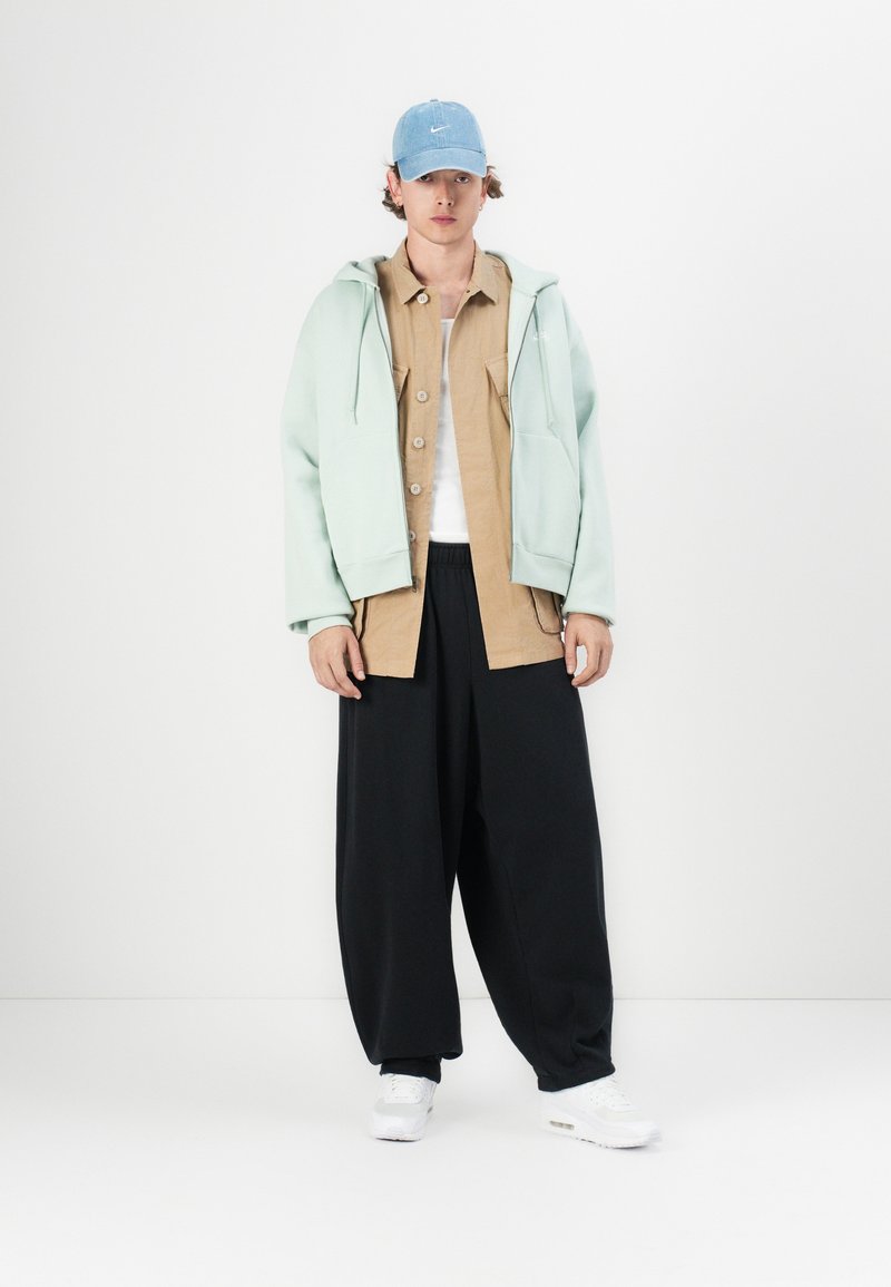 Jasno miętowy oversize'owy hoodie, brązowa koszula na guziki, czarne szerokie spodnie, białe sneakersy oraz jasnoniebieska czapka z logo.