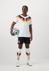 Άσπρη ποδοσφαιρική φανέλα με κόκκινες και κίτρινες διαγώνιες ρίγες, μαύρες λεπτομέρειες, μαύρη βερμούδα και καλτσάκια με σχέδιο που φέρουν το λογότυπο της DFB.