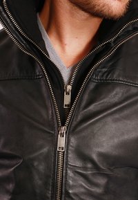 Svart läderjacka med en slät yta och silverfärgad dragkedja; har en huva och en grå tröja i lager under.