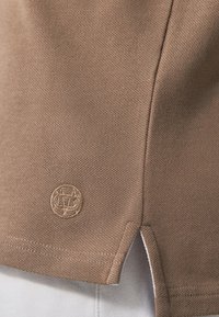 Brązowa bawełniana koszulka polo z fakturowanym materiałem, bocznymi rozcięciami i małym haftowanym logo w dolnym lewym rogu.