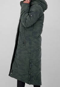 Grön kamouflagepuffjacka med huva, dragkedja och tryckknappar, som har ett texturerat tyg och sidofickor för extra funktionalitet.