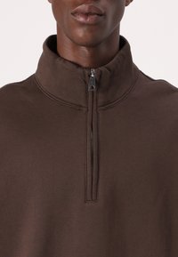 Brun pullover med høj krave og lynlås ved halsen. Fremstillet af et blødt materiale med glat tekstur, der har subtile syningsdetaljer.