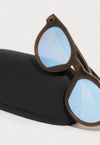 Aurinkolasit, joissa on ruskeat muovikehykset ja sinisävytetty linssi, lepäävät mustalla kovalla kotelolla, jossa on teksturoitu pinta.