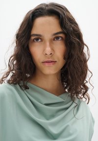 Top drappato verde chiaro realizzato in tessuto morbido, con scollo a cappuccio e superficie liscia, con capelli scuri ricci parzialmente visibili.