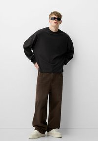 Crni oversize dukserica s dugim rukavima, uparen s smešnim hlačama širokih nogavica i belim patikama, postavljenim na jednostavnoj beloj pozadini.