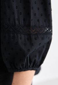 Black fabric with textured dot pattern, featuring a lace trim near the elbow. Soft, gathered sleeves with an elastic finish at the cuff.