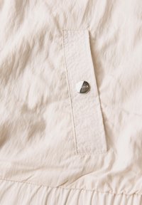 Beige stof met een kreukelige textuur heeft een zijvak met een zilveren drukknoop. Bevat zichtbare stiksels langs de randen.