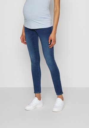 Personne enceinte portant un haut bleu clair, un jean de maternité bleu foncé et des baskets blanches, debout devant un fond uni clair.