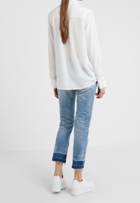 Blusa blanca de manga larga con canesú en la parte trasera, combinada con jeans ajustados azul claro con dobladillos deshilachados y zapatillas blancas.