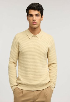 Jeune homme aux cheveux bouclés foncés, portant un pull polo beige texturé à manches longues et un pantalon beige, mains dans les poches, fond neutre.