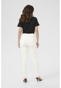 Jean blanc slim, poches arrière, taille haute, associé à un t-shirt noir. Le modèle porte des sandales à talons ouverts.