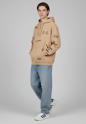 Felpa con cappuccio in beige chiaro, con testo grafico e design in rosso e nero. Abbinata a jeans in denim blu chiaro e sneakers bianche.
