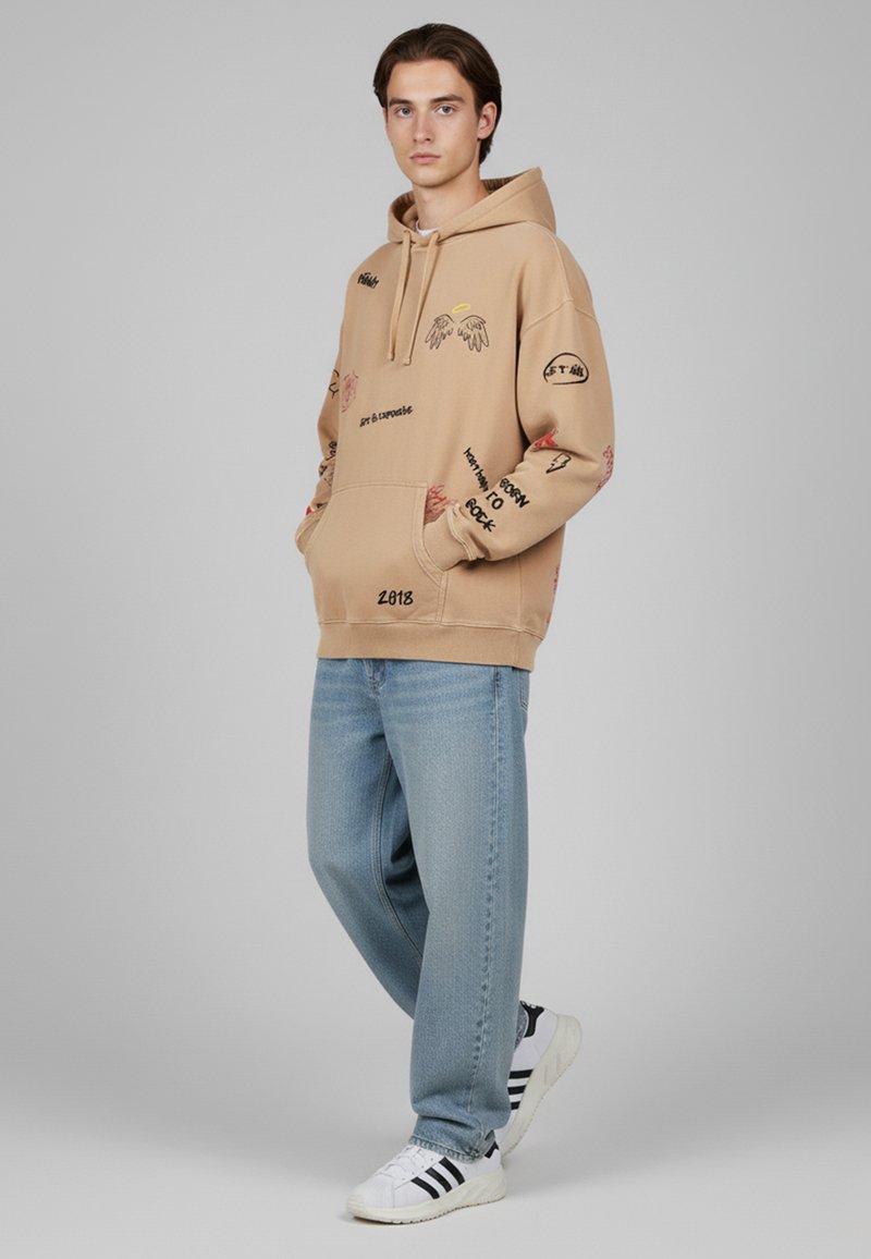 Sweat à capuche beige clair, présentant des textes graphiques et des motifs en rouge et noir. Assorti à un jean en denim bleu clair et des baskets blanches.