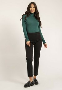 Pull à col roulé vert, pantalons noirs ajustés et chaussures à plateforme noires. La tenue présente une texture lisse et des éléments de design simples.