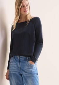 Marineblaue Strickjacke mit langen Ärmeln und weitem Rundhalsausschnitt, kombiniert mit hellblauen Jeans. Die Strickjacke hat gerippte Bündchen und eine glatte Textur.