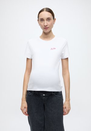 T-shirt en coton blanc à manches courtes, col rond, avec un petit graphique rose sur le côté gauche de la poitrine. Associé à un pantalon en denim foncé.