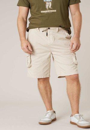Homme portant un short cargo beige, des baskets blanches et un t-shirt vert olive avec un motif d'ours et du texte à l'avant.