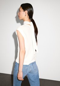 Beige sleeveless top with a tied neck detail, paired with light blue jeans. The background is plain white, highlighting the outfit.