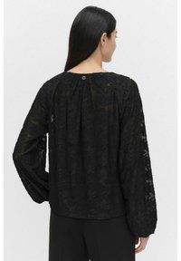 Svart blus med långa, genomskinliga ärmar och blommönsterbroderi. Har en rundad halsringning och en liten metallaccent på baksidan.