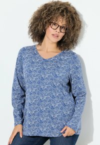 Donna con capelli ricci e occhiali, che indossa una maglietta manica lunga a fiori blu e jeans scuri, in posa su uno sfondo bianco.