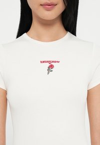 T-shirt blanc à fines côtes avec des manches courtes, ornée d'un texte "tommy jeans" brodé en rouge et d'un motif de rose sur la poitrine.