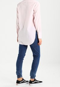Camicia a maniche lunghe rosa chiaro con orlo curvo, abbinata a jeans blu con risvolto e sneaker nere con dettagli bianchi.