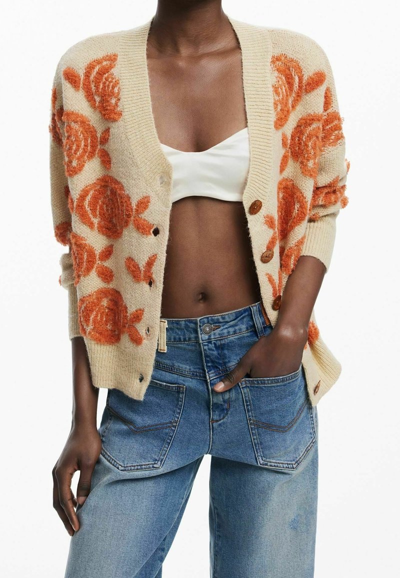 Personne portant un cardigan beige avec des motifs floraux orange, un crop top blanc et un jean bleu taille haute avec une main dans la poche.