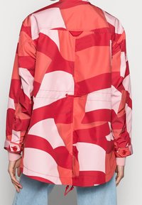 Rød og pink mønstret jakke med afslappet pasform, med høj krave, to knapper ved ærmekanterne og en taljeret midje med snøre.