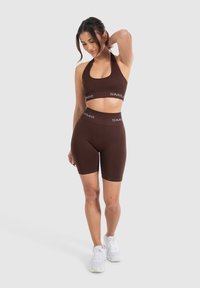 Reggiseno sportivo marrone e pantaloni bike a vita alta abbinati, con logo "SMILE" in bianco. Tessuto liscio e elastico. Modella in posa con postura rilassata.