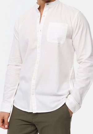 Homme portant une chemise blanche à manches longues boutonnée avec une poche poitrine et un pantalon vert olive, mains près des poches.