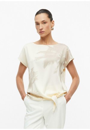 Femme aux cheveux foncés portant un chemisier crème à manches courtes avec un imprimé subtil de palmiers et un détail nœud, avec un pantalon blanc et des boucles d'oreilles créoles dorées.