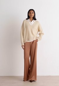 Light beige knitted sweater with a V-neck, paired with wide-leg brown trousers in a smooth fabric, creating a relaxed, layered look.
