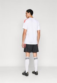 Λευκό αθλητικό φανελάκι με κόκκινες ρίγες στους ώμους, μαύρο σορτς, λευκές κάλτσες μέχρι το γόνατο με μαύρες ρίγες και μαύρα ποδοσφαιρικά παπούτσια. Πλάτη.