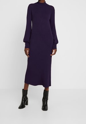 Femme portant une robe midi en maille violette à manches longues, col montant, manches retroussées et bottines à talons noires sur fond blanc.