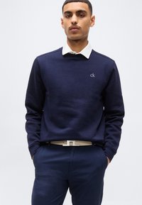Homme portant un pull bleu marine avec une chemise à col blanc en dessous, un pantalon bleu marine et une ceinture blanche, debout les mains dans les poches.