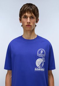 Blue cotton t-shirt featuring white graphic prints and logos on the front. Short sleeves, round neckline, and a relaxed fit design.