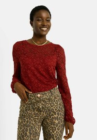 Haut en dentelle rouge à manches longues avec des motifs en vagues, associé à un pantalon imprimé léopard. Accents en métal doré sur le pantalon.