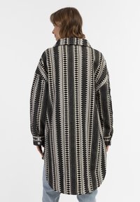 Didesnio juodai baltas marškiniai su apykakle, ilgomis rankovėmis ir suapvalintu sijonu, pagaminti iš tekstūruotos medžiagos su vertikaliomis juostelėmis.