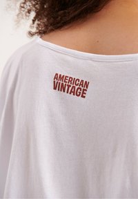 Balta medvilninė marškinė su laisvu kirpimu, su raudonu tekstu "AMERICAN VINTAGE" nugarinėje apykaklėje. Lygi tekstūra.